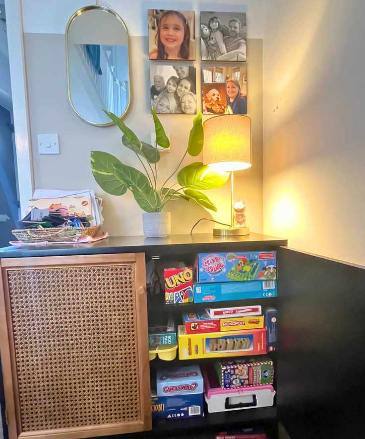 A black sideboard with rattan fronting open with three shelves of colorful board games showing. There is a lamp, plant and key box on top with pictures and a gold mirror above