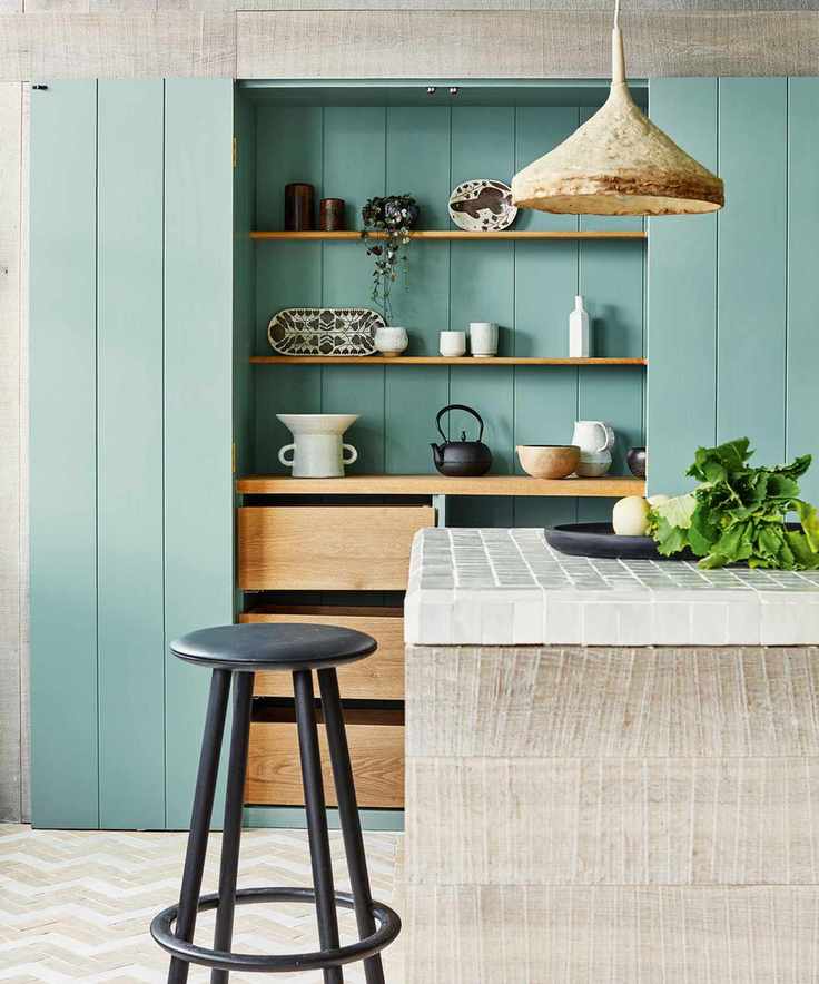 Kitchen with central island with a white tiled countertop, timber clad wall and unit, green and wood cupboards and black stool.
