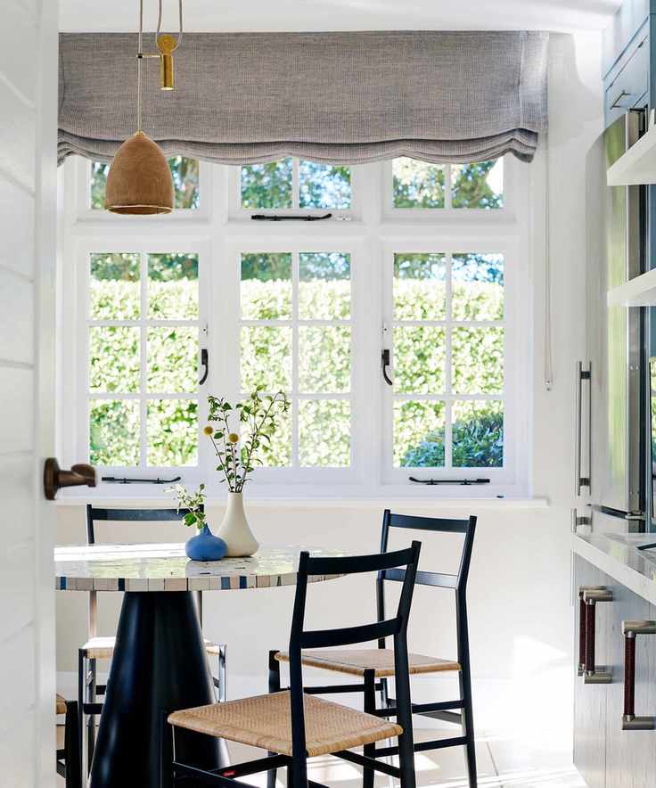 A large white framed window in a dining room, with a grey fabric roman blind across the top. A marble round dining table in front of it surrounded by black chairs with woven wicker bases.