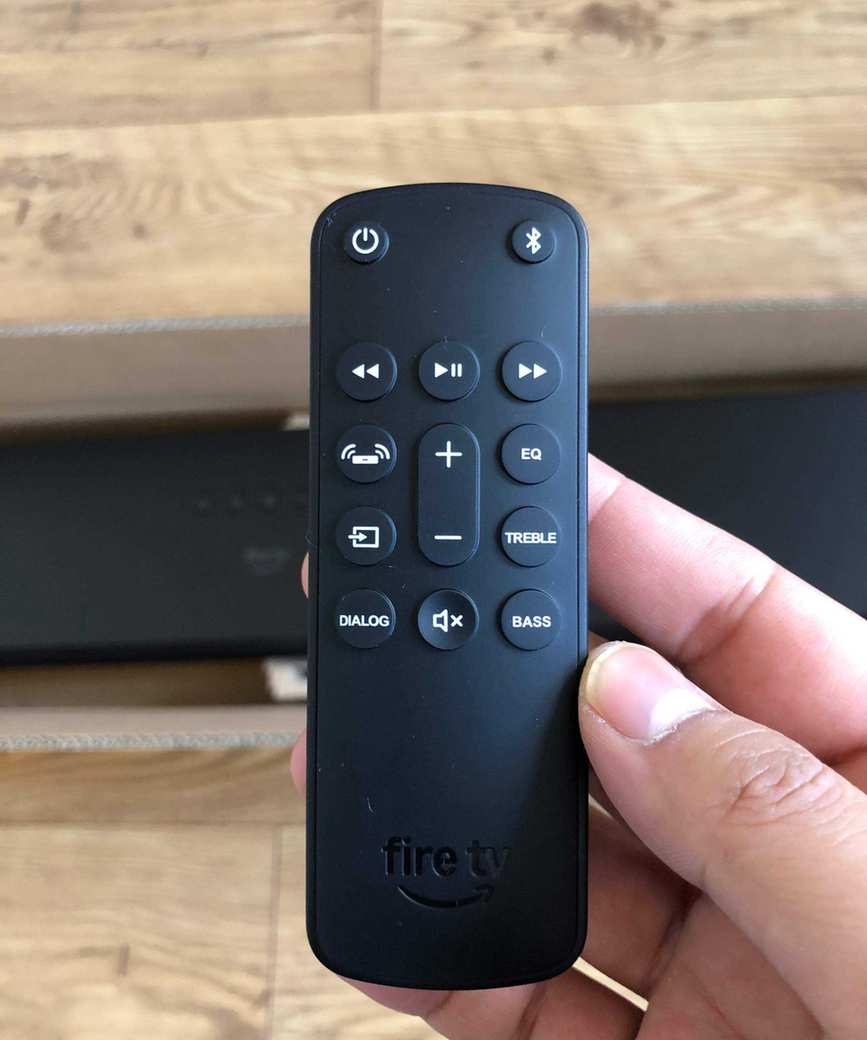 Remote for the Fire TV Soundbar Plus on a wooden floor with the soundbar visible.