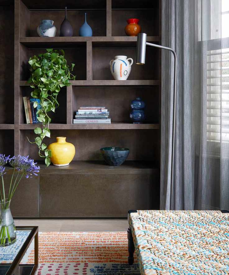 living room with brown custom storage with shelves, gray voile drapes in front of shutters, woven seat, woven rug, coffee table, plants, flowers, floor lamp, vases and ornaments
