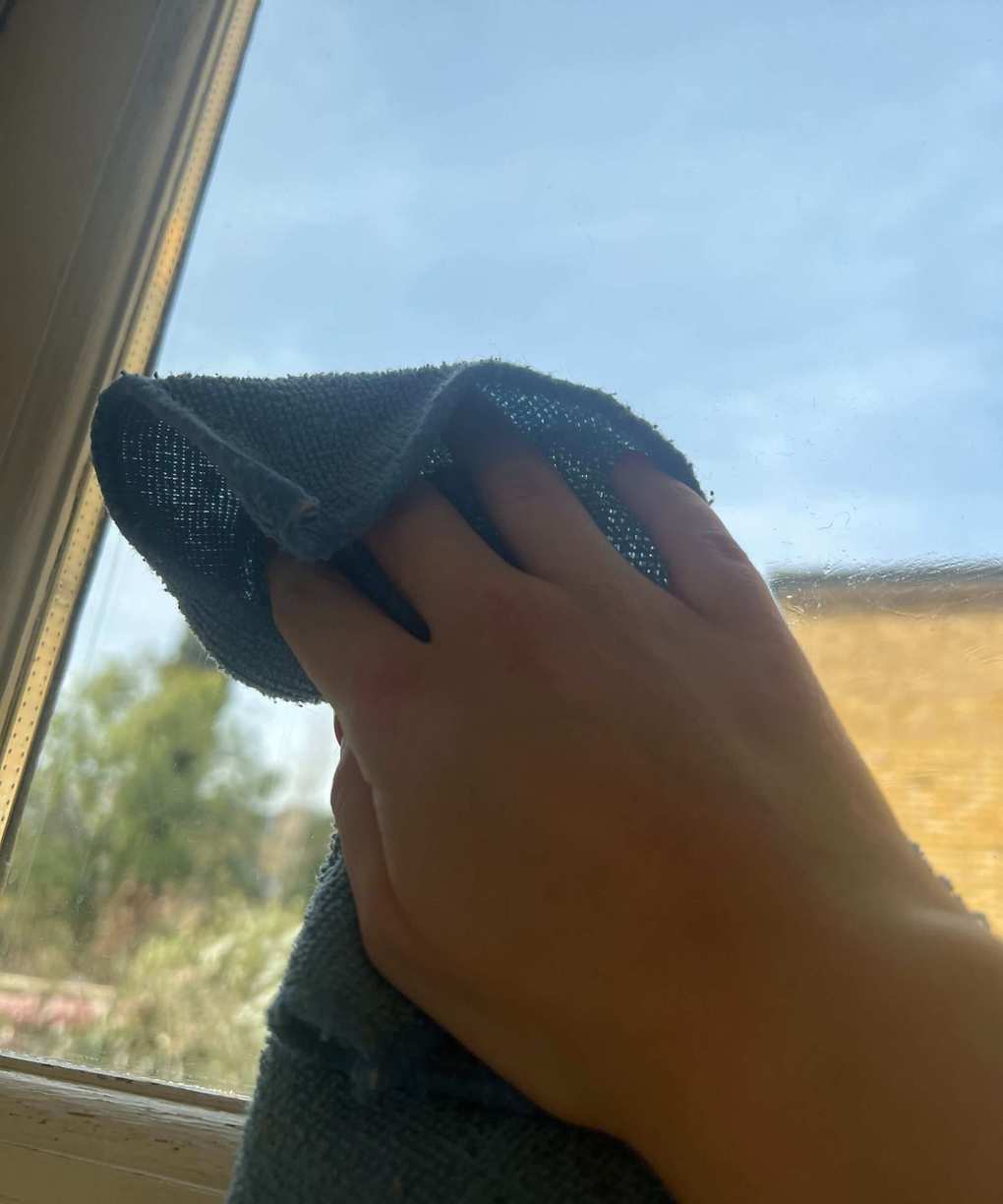 A hand wiping a window with a blue microfiber cloth, with sunny blue skies behind