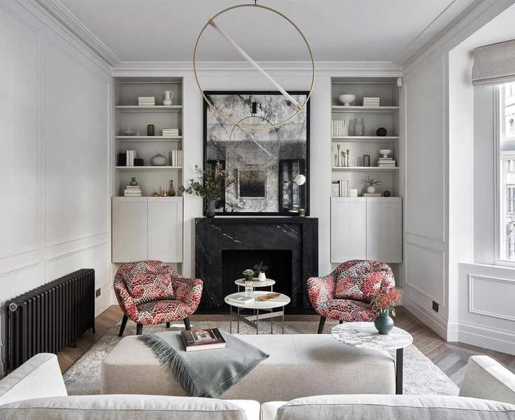 Living room with pale grey walls, bay window, built in storage either side of the chimney breast and fireplace. Grey rug and sofa and orange patterned armchairs.