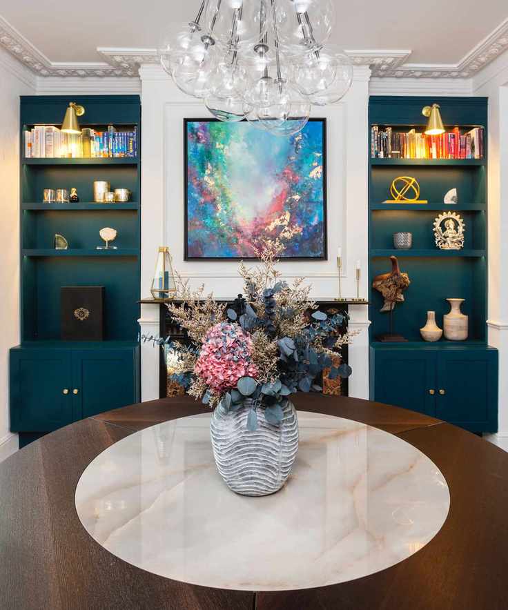 dining room with round mahogany table, vase of flowers, teal matching custom storage in alcoves each side of fireplace, artwork above fireplace, globe chandelier