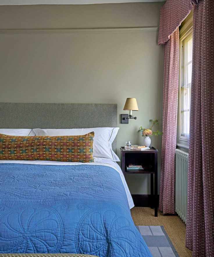 Bedroom with sage green walls, grey fabric headboard, blue quilt and long colorful bolster, rattan carpet and pink pelmet curtains around the window