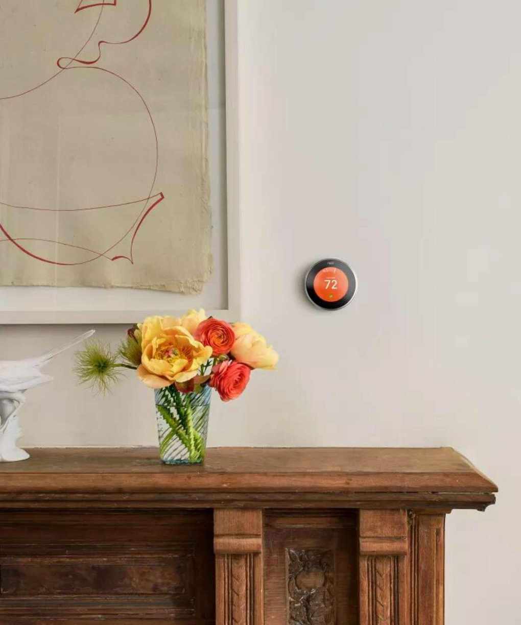 A small orange smart thermostat on a beige wall above a brown sood side boared with red and yellow flowers