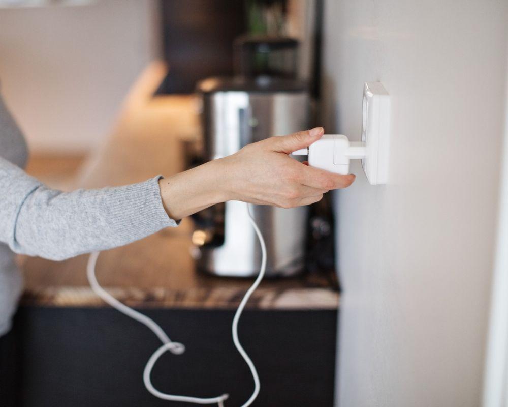 A person wearing a gray jumper plugging in a white plug, with a kitchen counter in the background