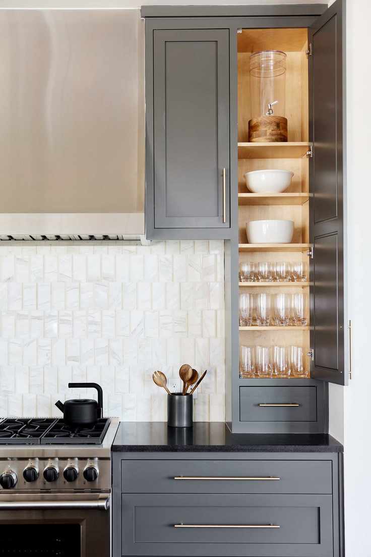 A grey kitchen with a tall cabinet full of glasswear beside a gas stove