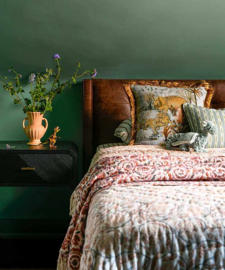 A bedroom featuring deep green walls, patterned bedding, and a stylish nightstand with flowers.