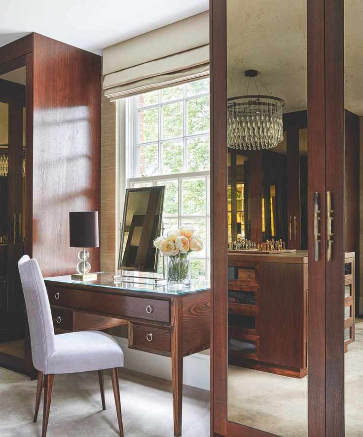 Mirrored brown closets in bedroom with a brown mirrored dressing table, white chair, mirror and large bright window with beige blinds.