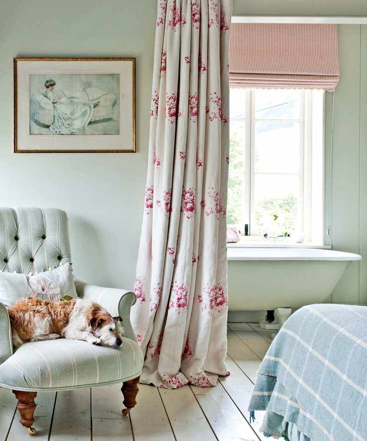 Neutral bedroom and bathroom, separated by floral curtain, roll top bath, pet dog on upholstered armchair, white painted floorboards