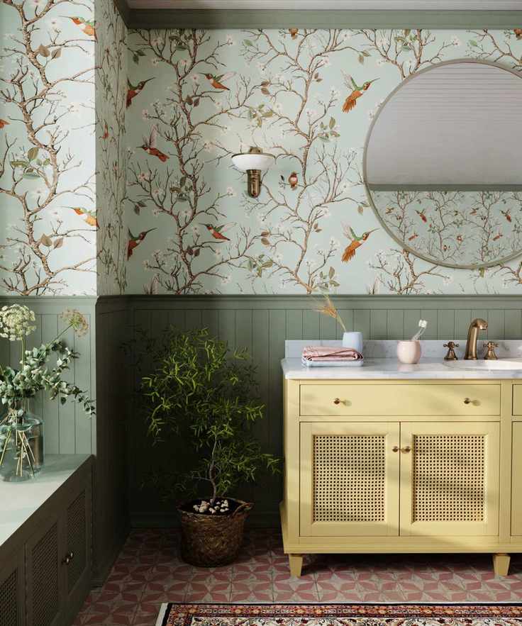 Sage green half paneled bathroom with butter yellow vanity and the Bronte tree and bird wallpaper. There are potted plants, a window seat, and tiled geometric orange floor tiles with a bathroom rug