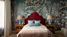 A bedroom with teal floral wallpaper, a burgundy headboard, blue and burgundy bedding, and two wooden nightstands with blue lamps.