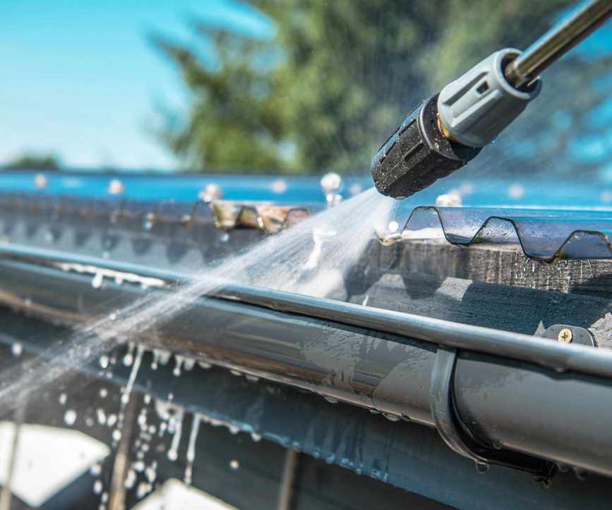 Close up of pressure washer washing a roof gutter with out-of-focus tree and sky in background.