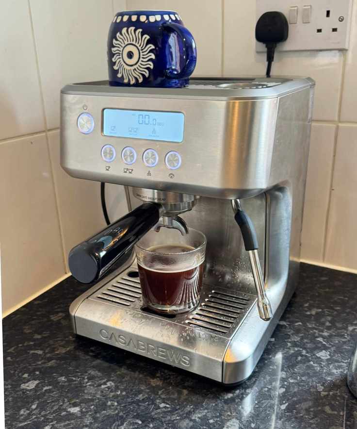 Casabrews ULTRA Espresso Machine on black countertop in front of white tiles, with blue mug positioned upside down on top of the coffee machine.