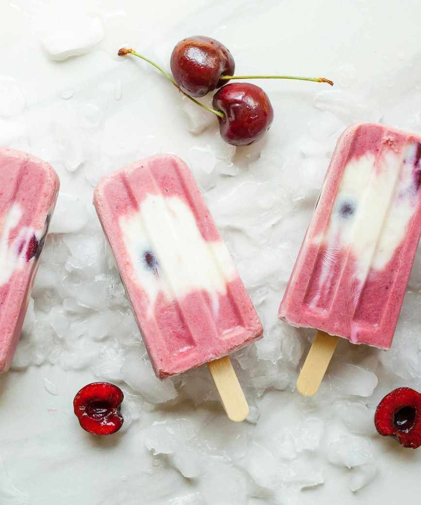 Cherry and vanilla popsicles on a white marble background, with pitted cherries scattered around them