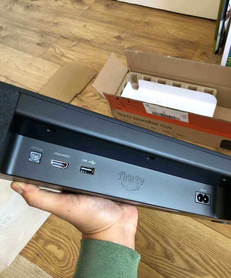 Hand holding the Fire TV Soundbar Plus, showing various ports, on a wooden floor.