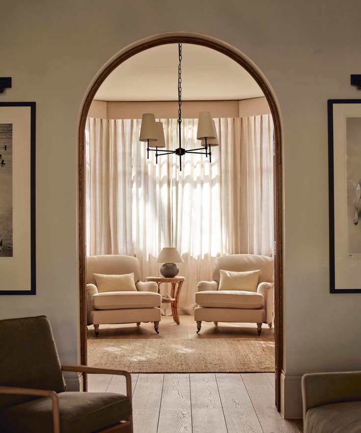 neutral living room with two armchairs and sheer curtains with an archway into the next room