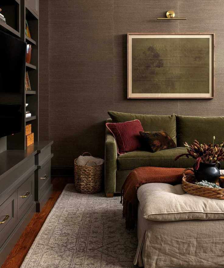 Dark living room with patterned rug, green sofa with cushions, framed minimalist painting, partial black bookshelf to the left, footstool, wooden floors.