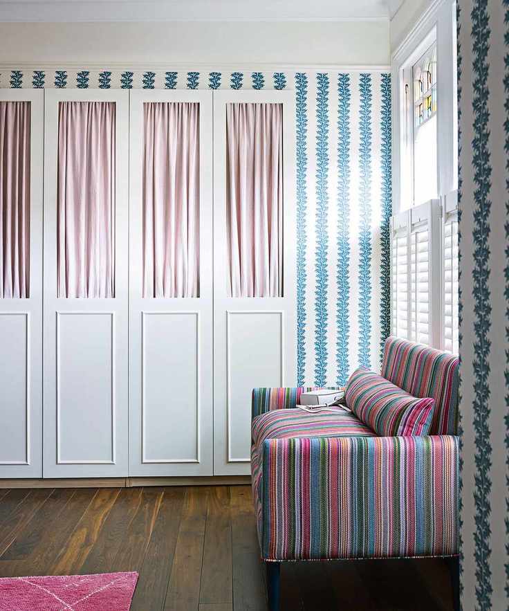 Bedroom with dark wooden floor colourful striped sofa, blue and white wallpaper and built in wardrobes, pink fabric curtains