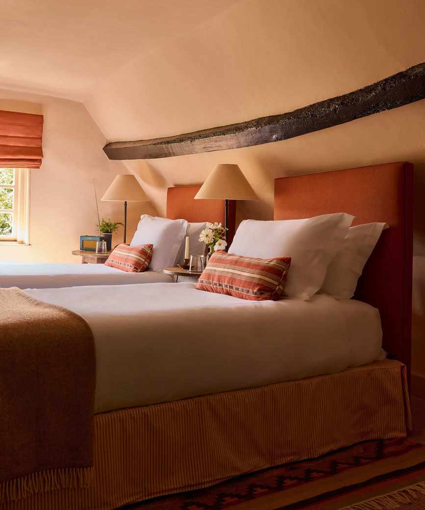 A loft bedroom with sloped ceilings color-drenched plaster pink, an exposed wood ceiling beam, and twin beds with terracotta headboards, crisp white bedding, and terracotta cushions and throws.