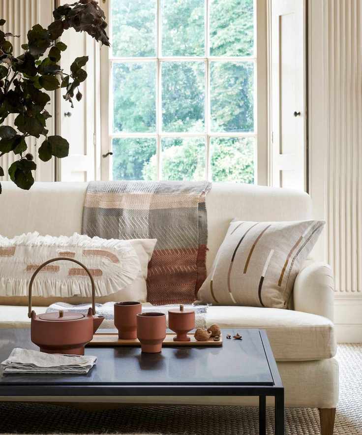 A white sofa with wooden legs in a white room, with sisal carpet, dark grey coffee table, cushions and blankets, a terracotta tea set on a wooden tray, and a dark green leafy plant