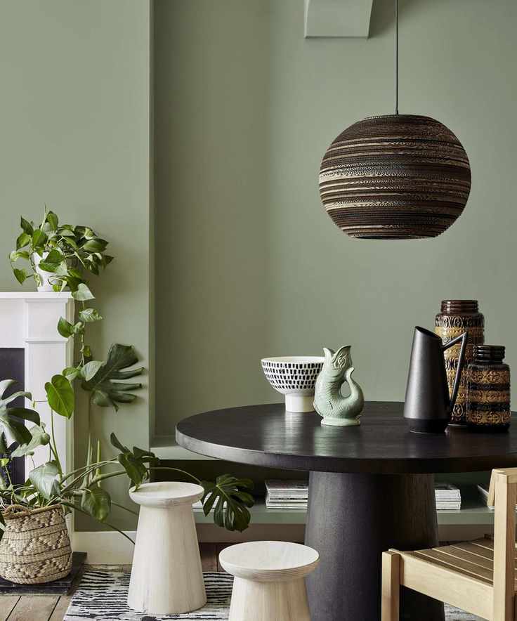 A matte green dining room with a round black dining table, wooden chairs, and a low hanging black pendant light