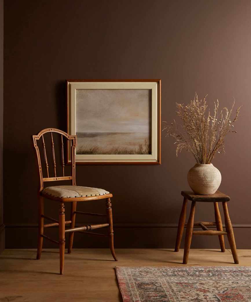 dark brown wall in living space with wooden chair, wooden stool with vase of dried stems and landscape framed picture on wall