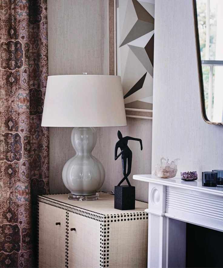 A living room with a grey lamp and shade on a small cupboard in an alcove beside a fireplace, and long patterned curtains.