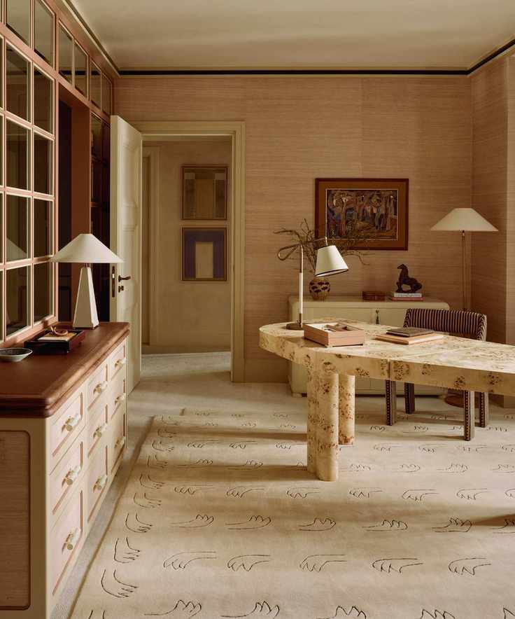 Neutral office space featuring an asymmetrical burl wood desk, linen-shaded table lamps, wall art, textured wallpaper, a figural motif beige rug, and a striped office chair