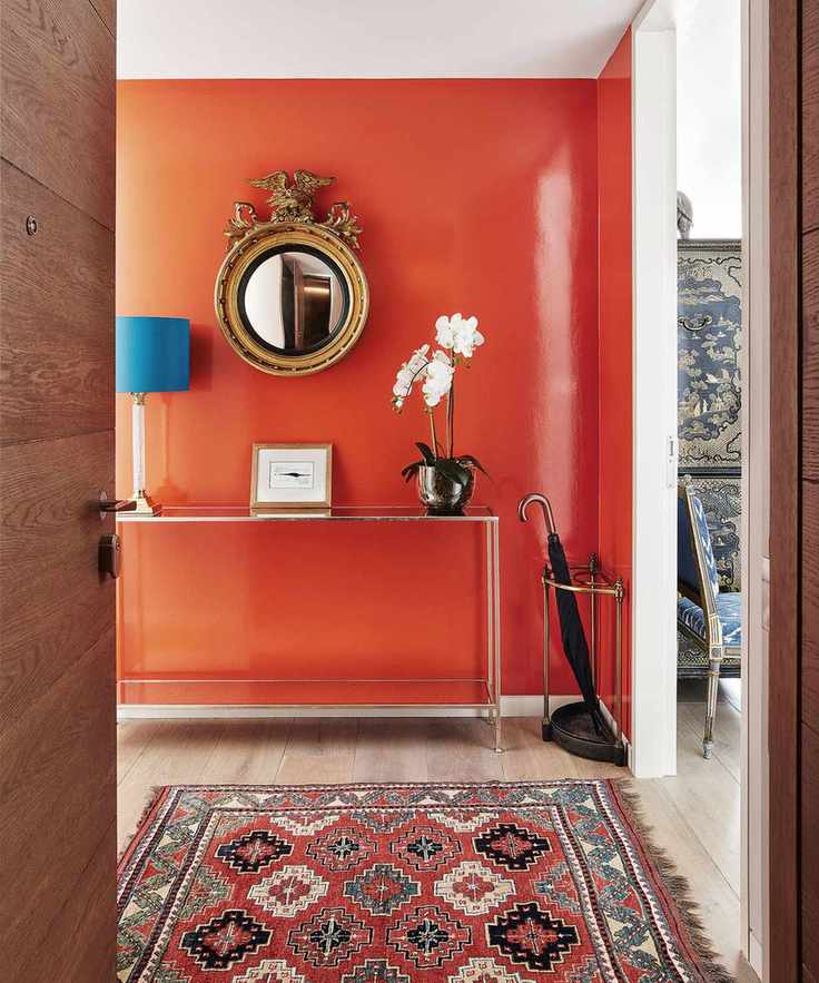 Hallway with a wall painted a bright orange with a matching rug in orange, metallic console table and umbrella stand, round mirror positioned above console table