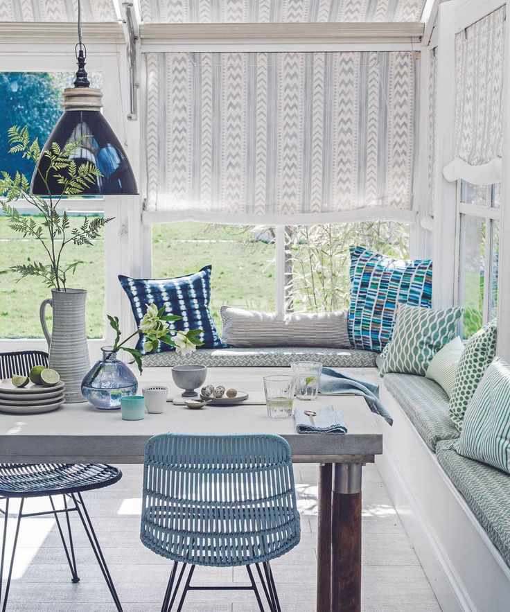A bright and sunny blue and white sunroom with green banquette seating and colorful, mismatched cushions. There is a large table laid with blue napkins, plates and vases, with a large white jug with a green leafy stem inside. Behind are large windows with white and grey patterned blinds half closed.