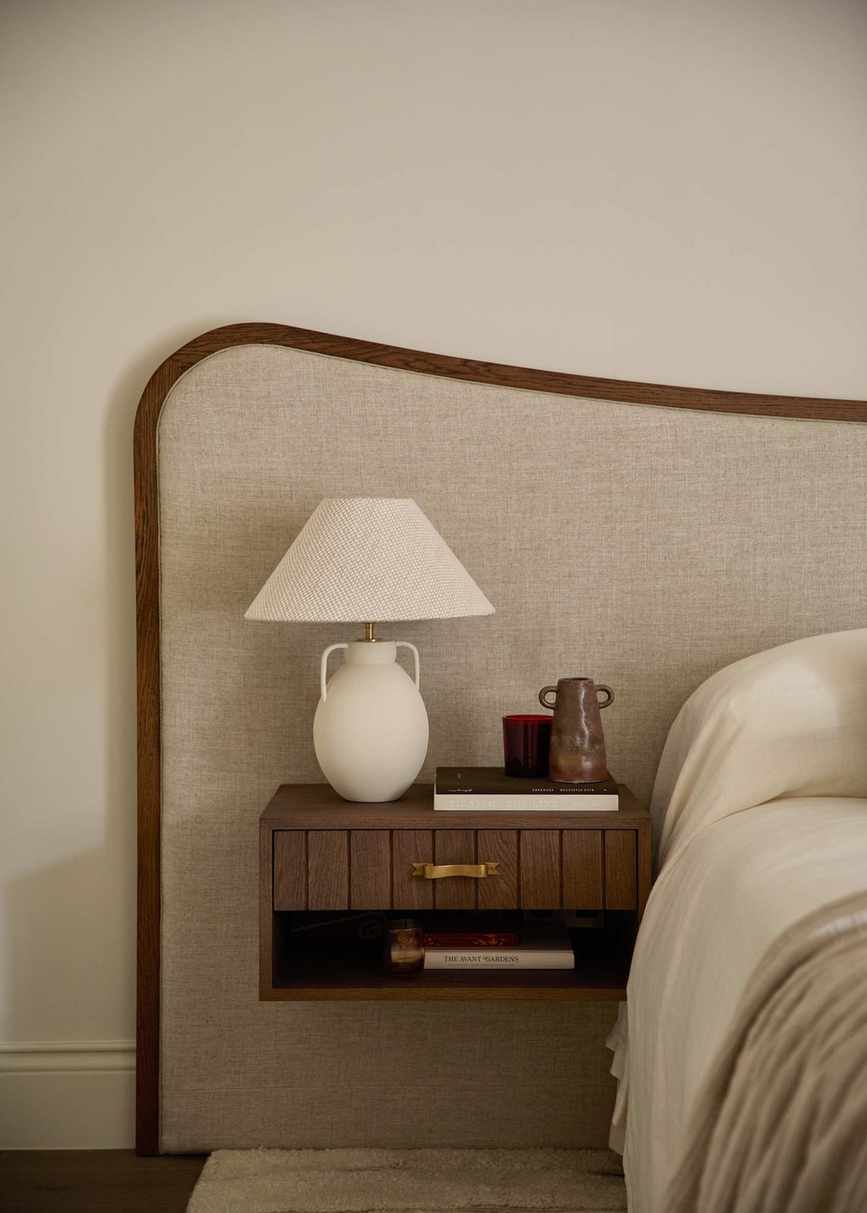 A bedroom with an oversized brown and taupe fabric headboard, a floating nightstand, and a neutral white lamp on the tabletop