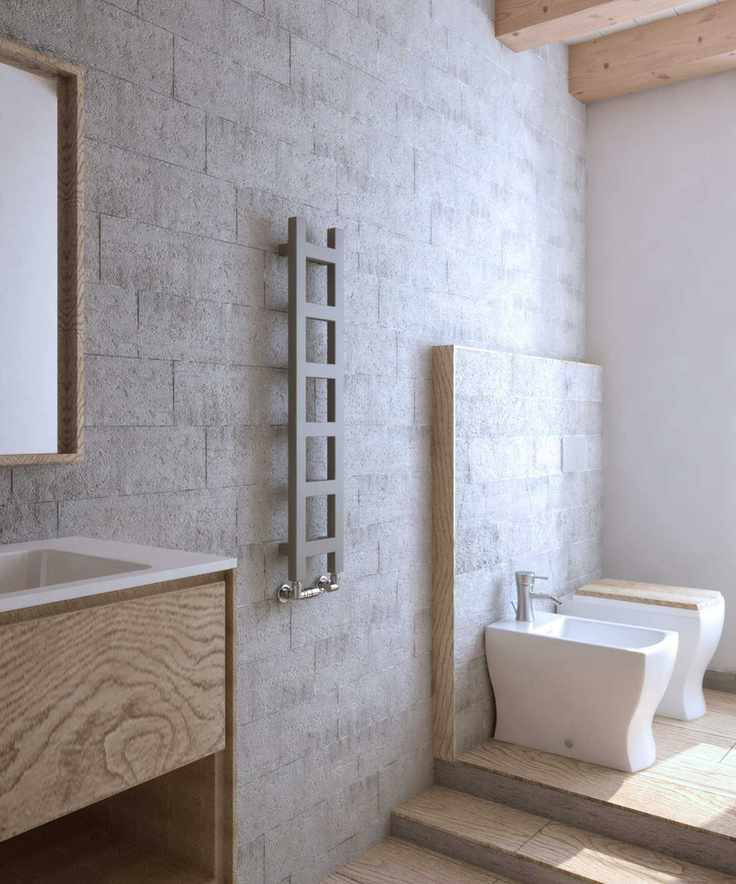 A gray ladder radiator on a stone bathroom wall, beside a wooden basin unit with a square mirror above, and some small steps up to a bidet and toilet.