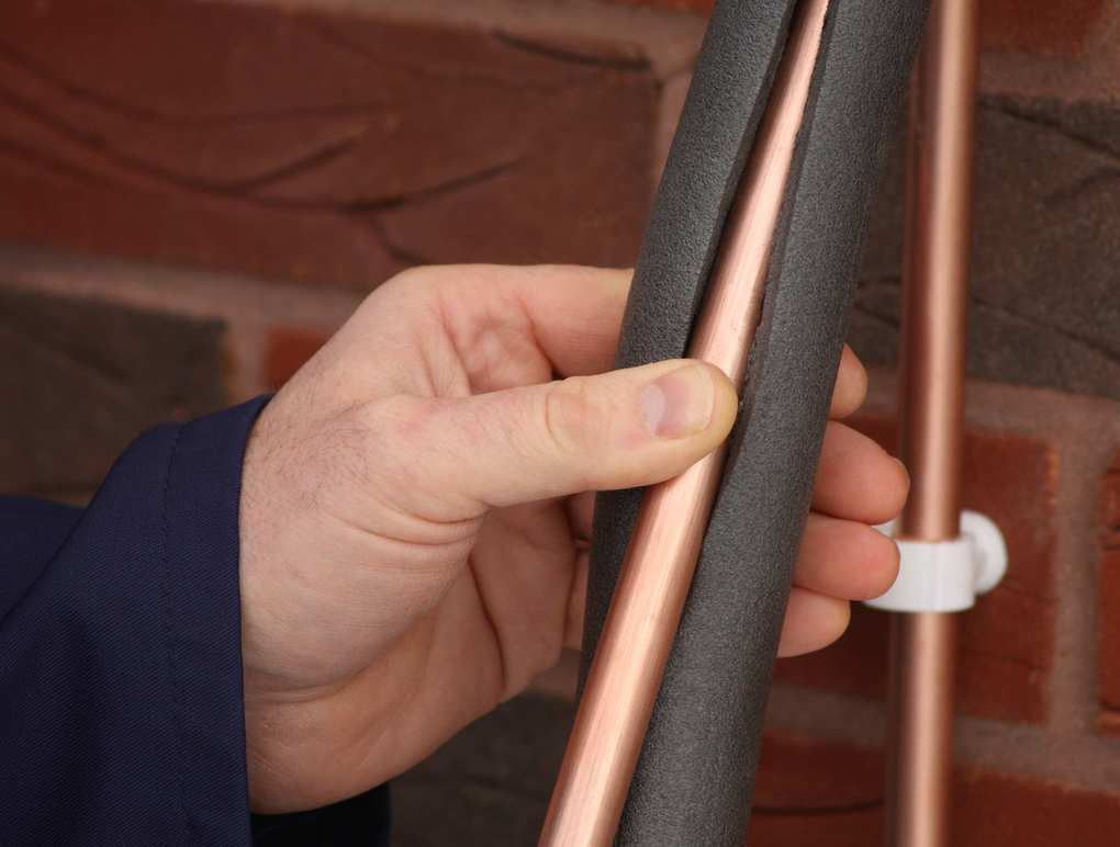 A man wrapping a copper pipe in a coat of foam insulation against a brick wall