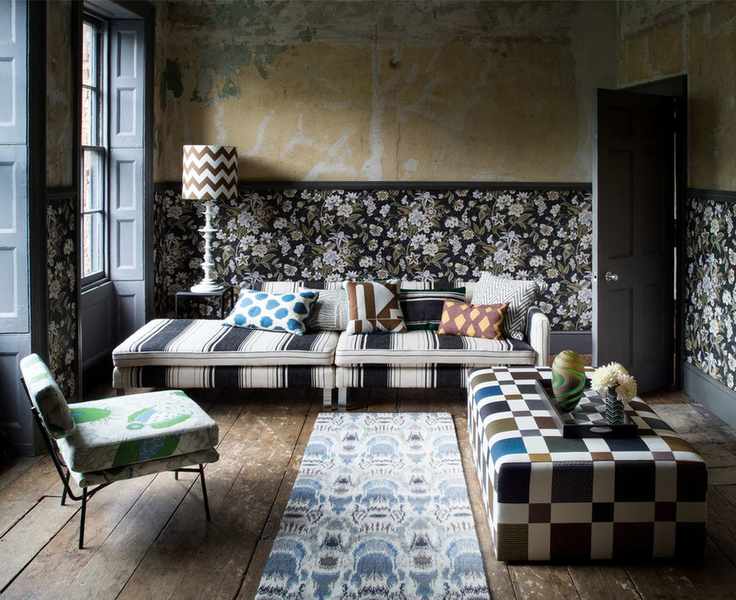 Living room with unfinished plaster walls and floral wallpaper, featuring a daybed sofa in black and white stripes and a checked metallic ottoman.