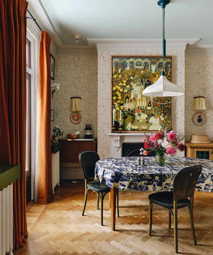 scandi maximalist style dining room with wallpapered walls, a large fireplace with folk art hung above, dark orange velvet curtains, a dining table with blue and white printed tablecloth and a white fabric pendant light