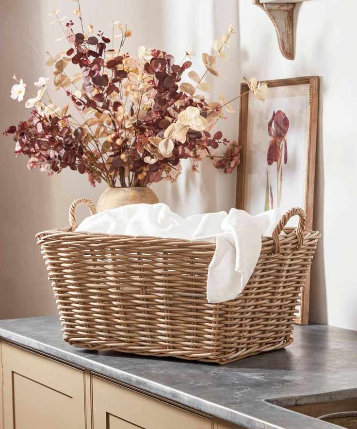 A small woven storage basket filled with a white sheet, on a grey counter. A vase of faux cream and pink stems behind it.