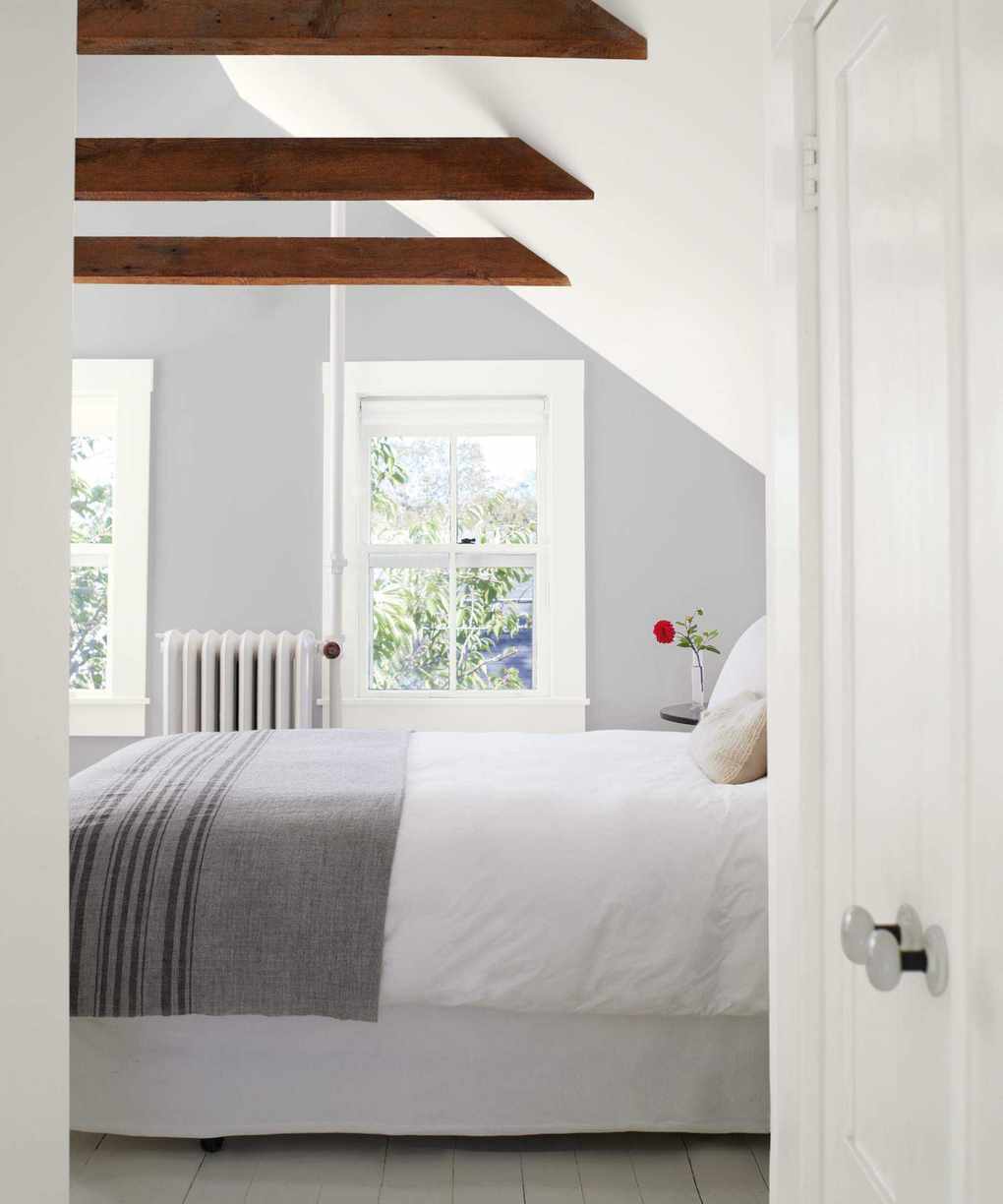 bedroom with light grey and white walls, grey blanket, beams, sloping ceiling, white floor bards, view of garden