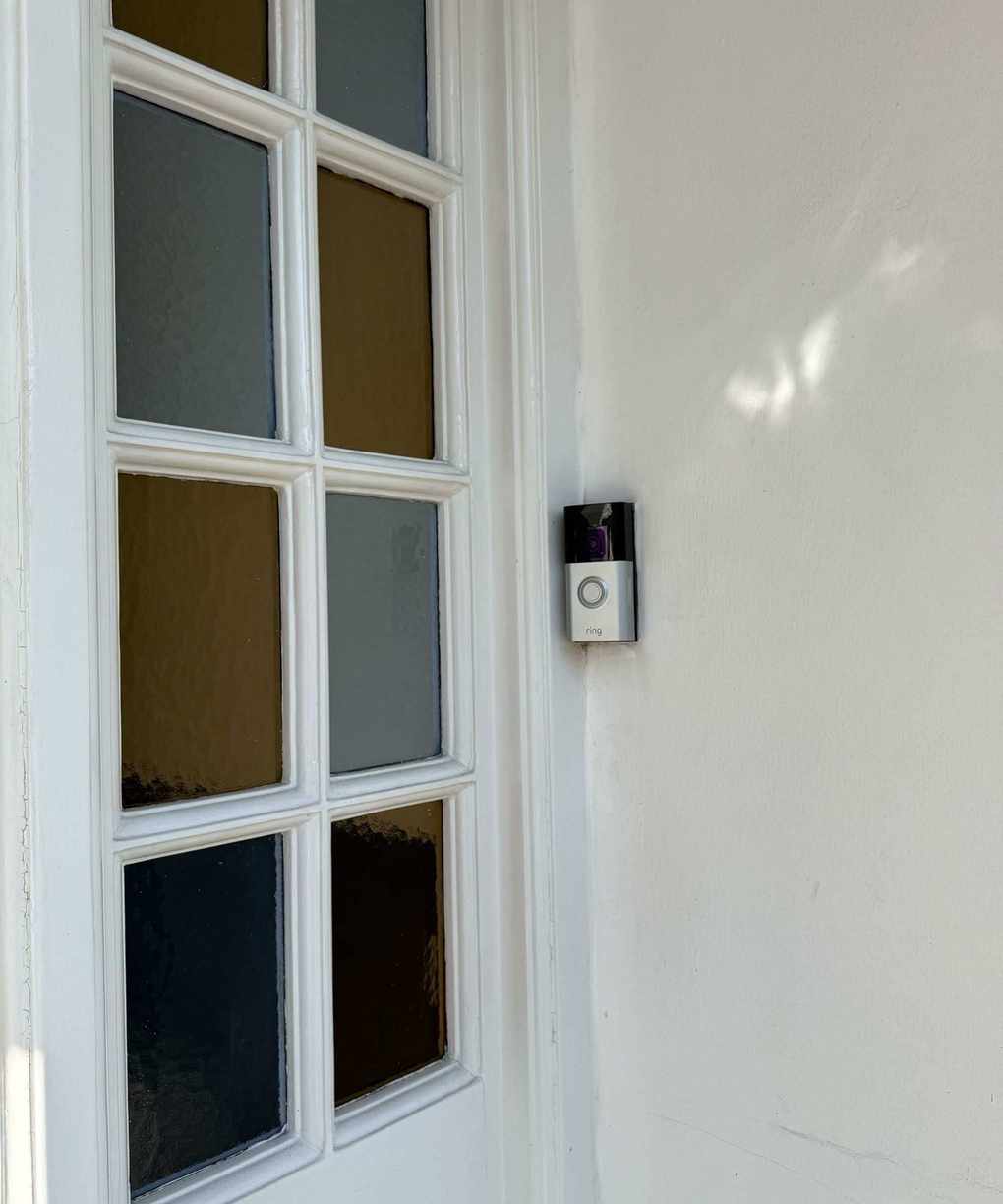 The Ring Battery Doorbell Plus mounted on a white painted wall, next to a yellow and blue panelled stain-glass window to the left of it.