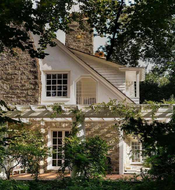 A traditional home exterior with exposed stone, white paint, and traditional-style windows, surrounded by greenery from trees.