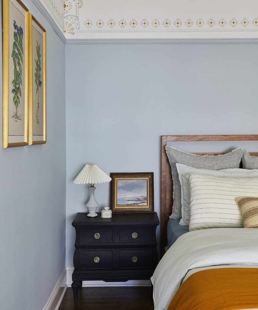 A pale blue bedroom with a black side table, lamp and painting