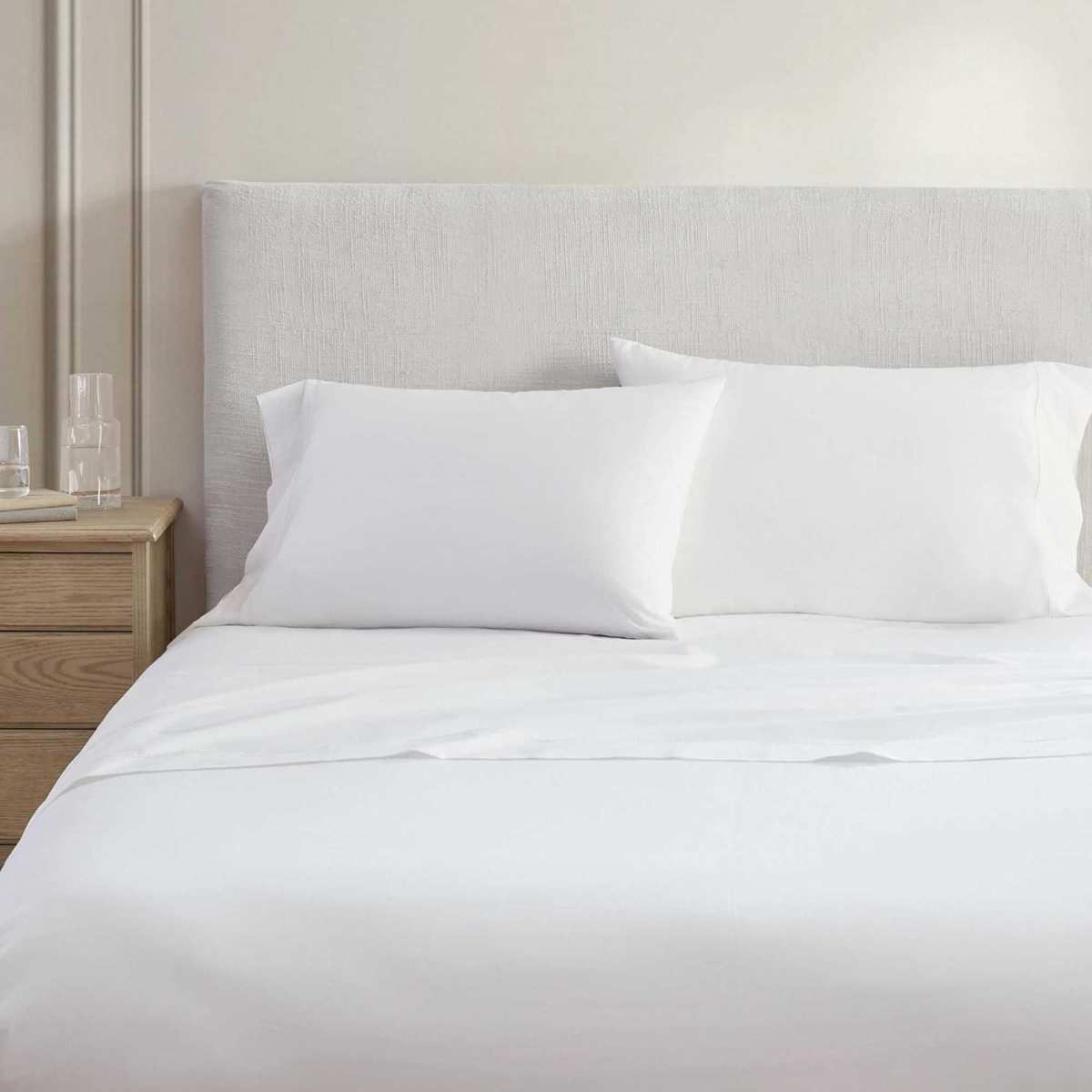 A white Martha Stewart Cotton Percale sheet set pictured on a pale white bed with a white panelled wall behind and wooden beside table with glasses to the left.