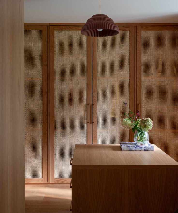 Built-in wall of wooden closets with rattan door fronts. A wooden island in the centre of he room with a small vase of flowers, and a rattan pendant light hanging overhead.