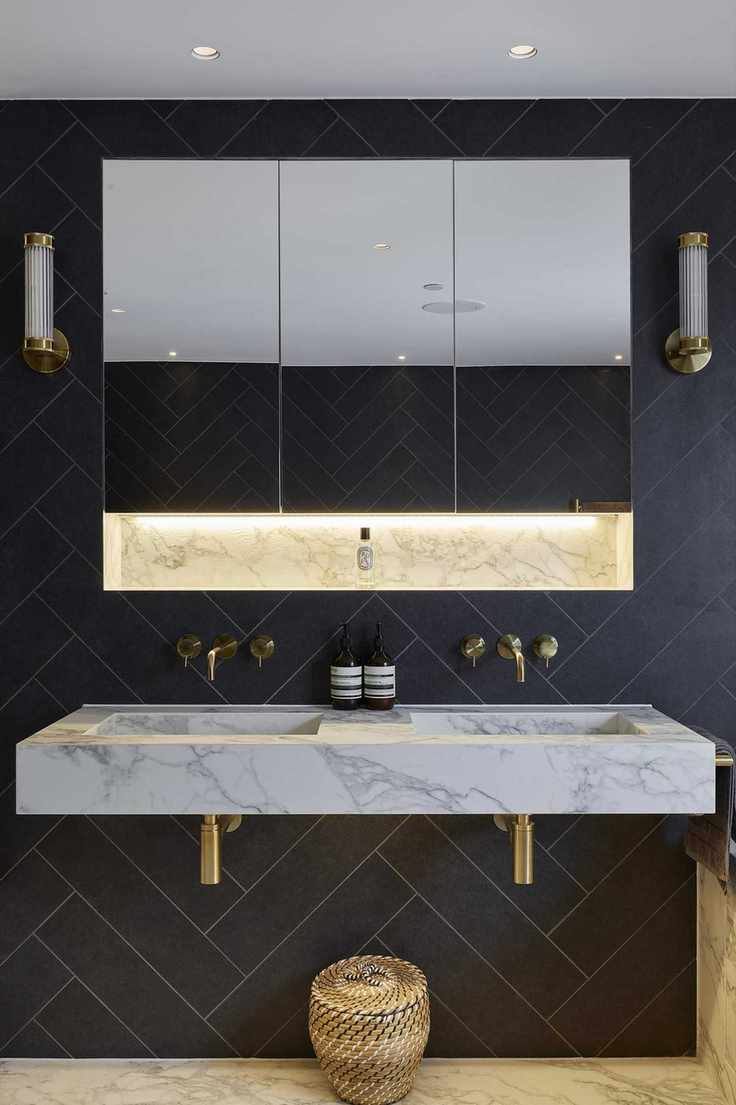 An example of dark bathroom ideas showing a marble basin with gold taps, gold wall lights and a mirror, and navy herringbone tiles