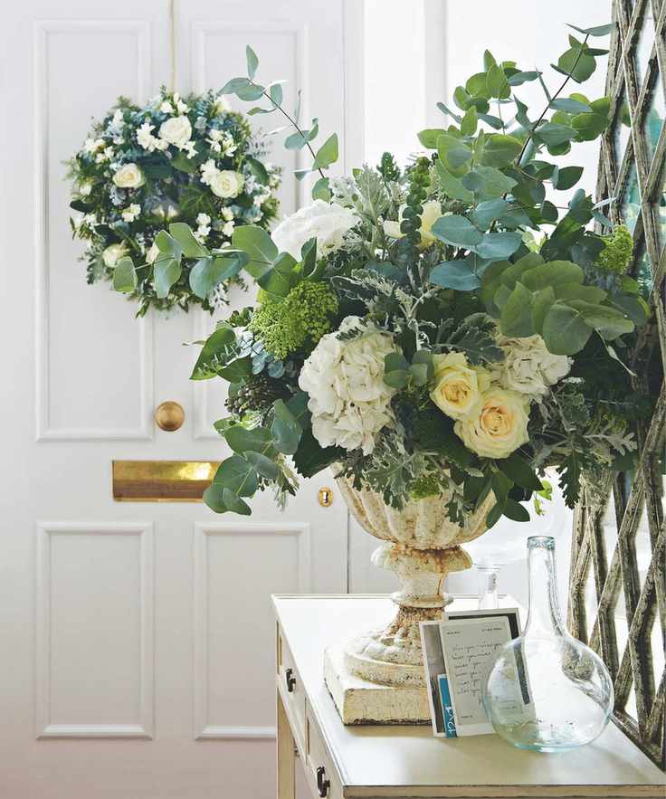 urn of white and green flowers and foliage on a hallway table with a white doorway in shot and a white and green wreath hanging on it