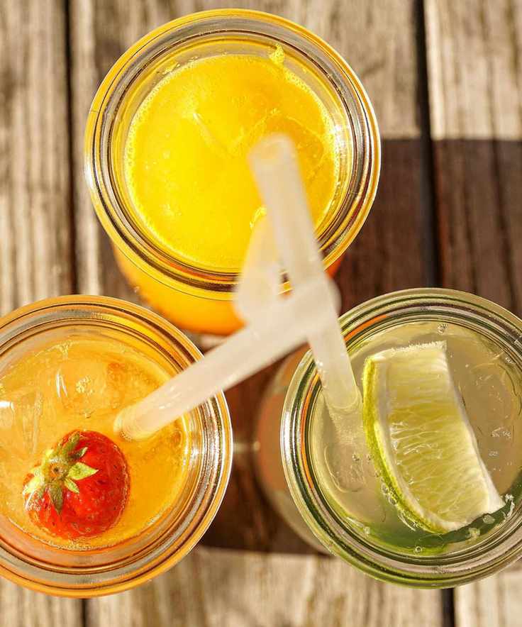 Aerial view of orange and lime cocktails served in mason jars with clear plastic straws, sitting on a wooden table