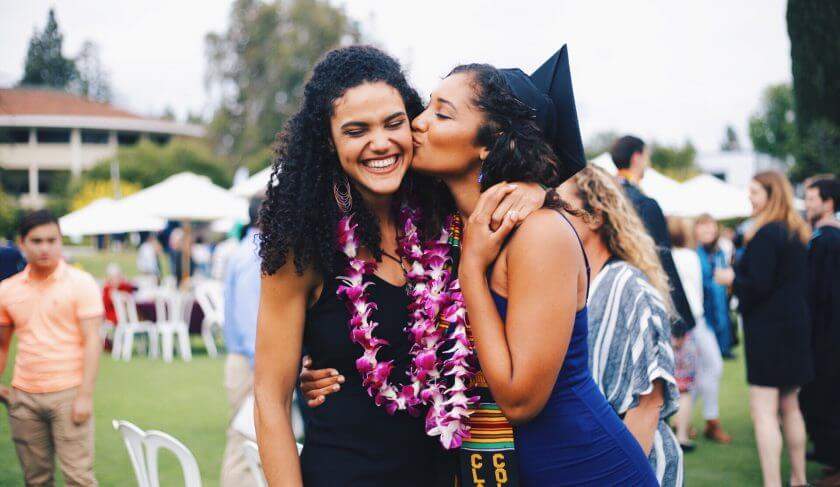 A Black woman and daughter are seen on the daughter's graduation day. The daughter is kissing the mother on the cheek.