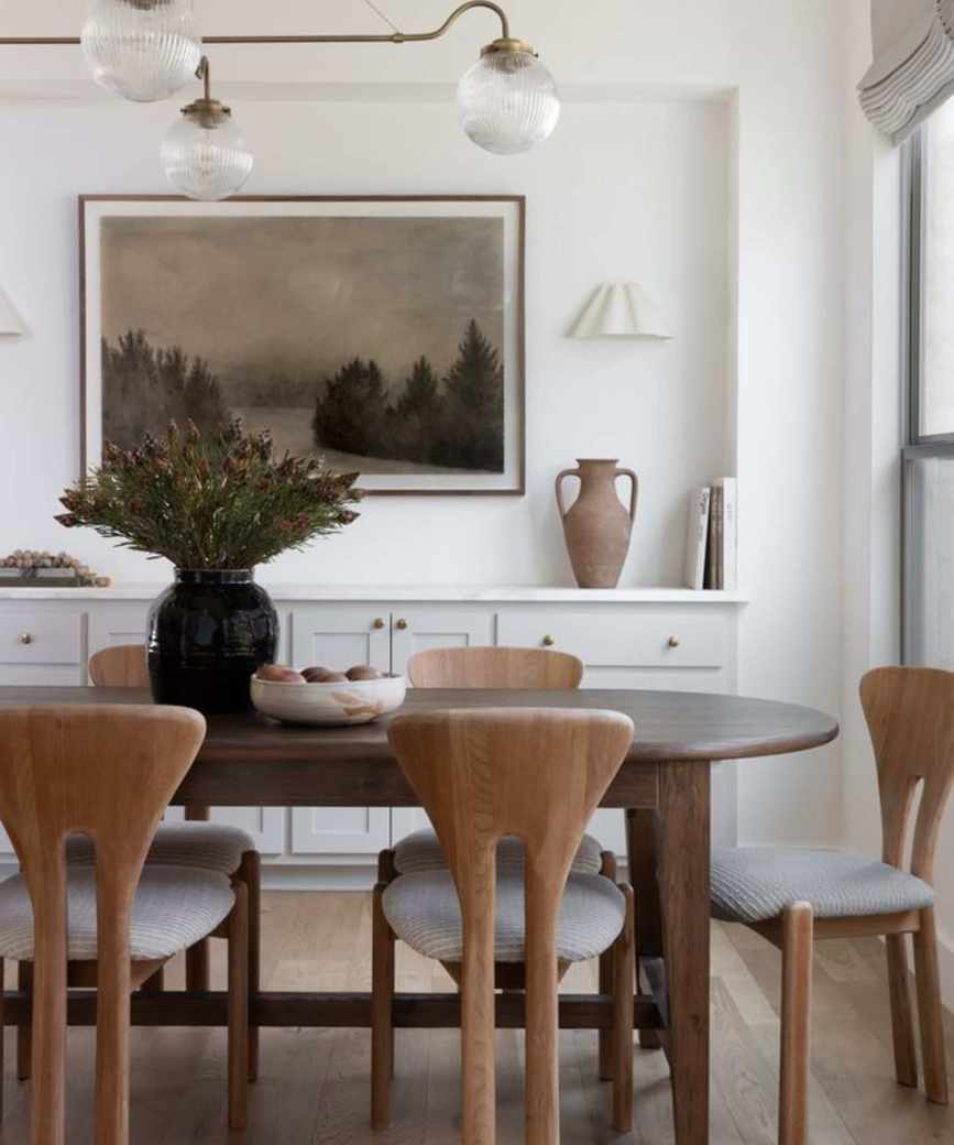 a white dining room with an oval wooden dining table, shapely wooden dining chairs, and brown accessories including artwork and a decorative jug