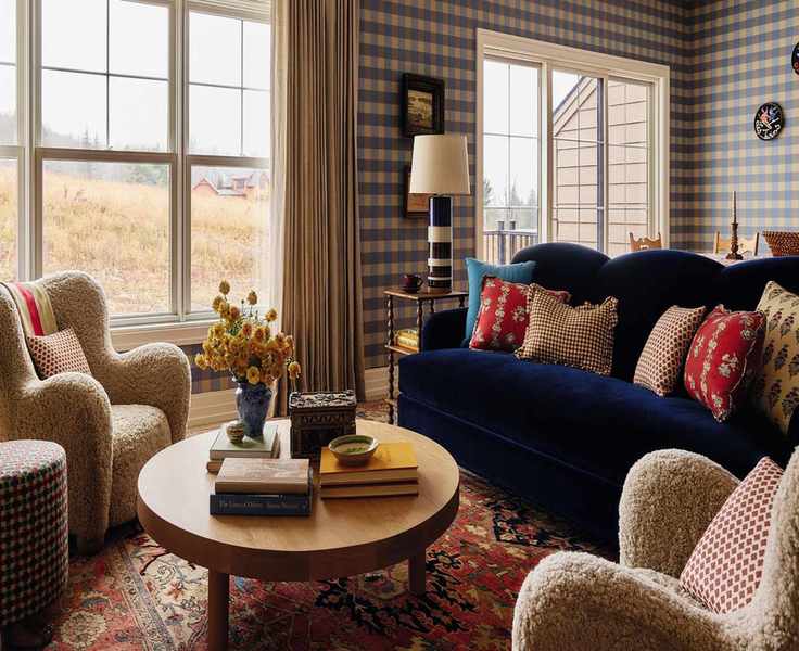 an open plan living area in a home in the Catskills with blue gingham wallpaper, a navy velvet couch, vintage red rug, sheepskin armchairs and a round wooden coffee table